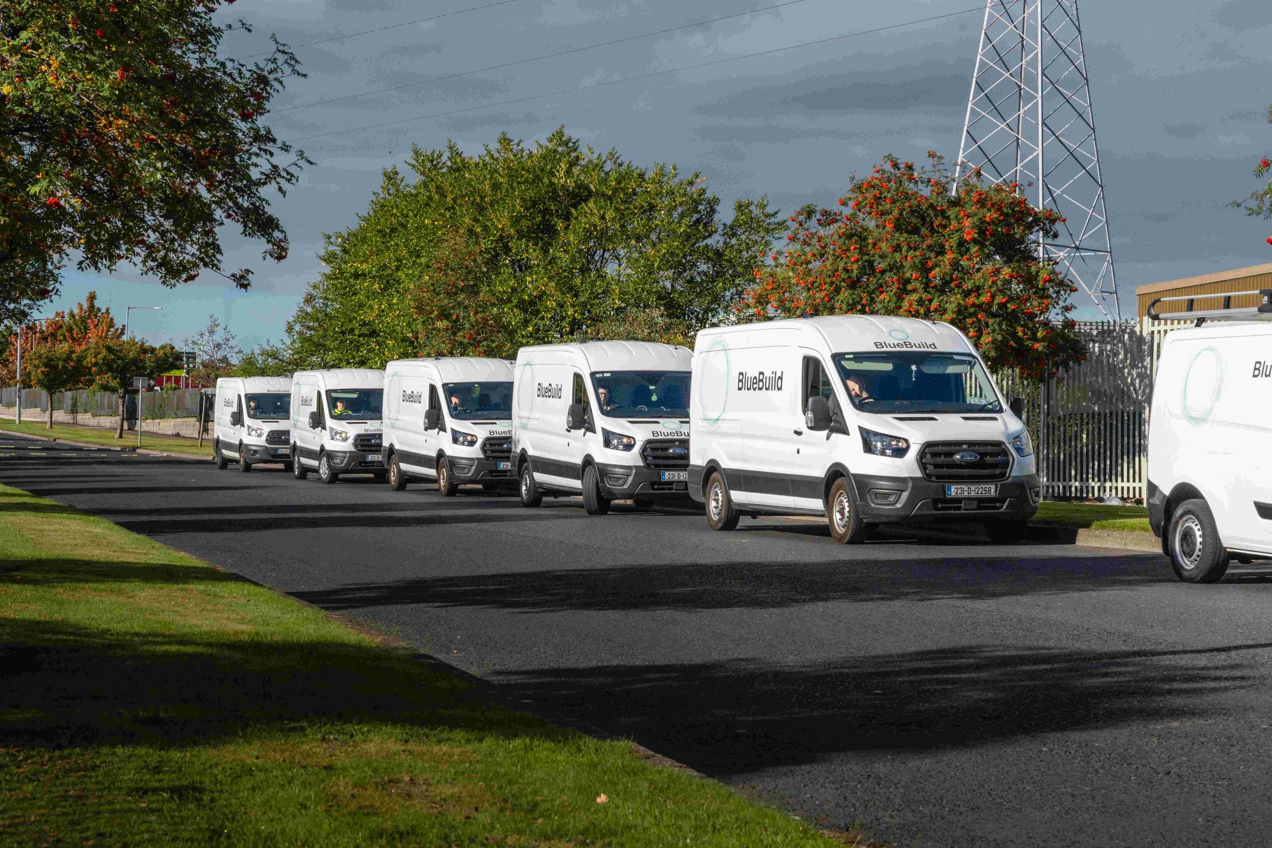 A fleet of branded white BlueBuild commercial transit vans driving in a convoy line along a public road with green trees and an electricity pylon in the background.