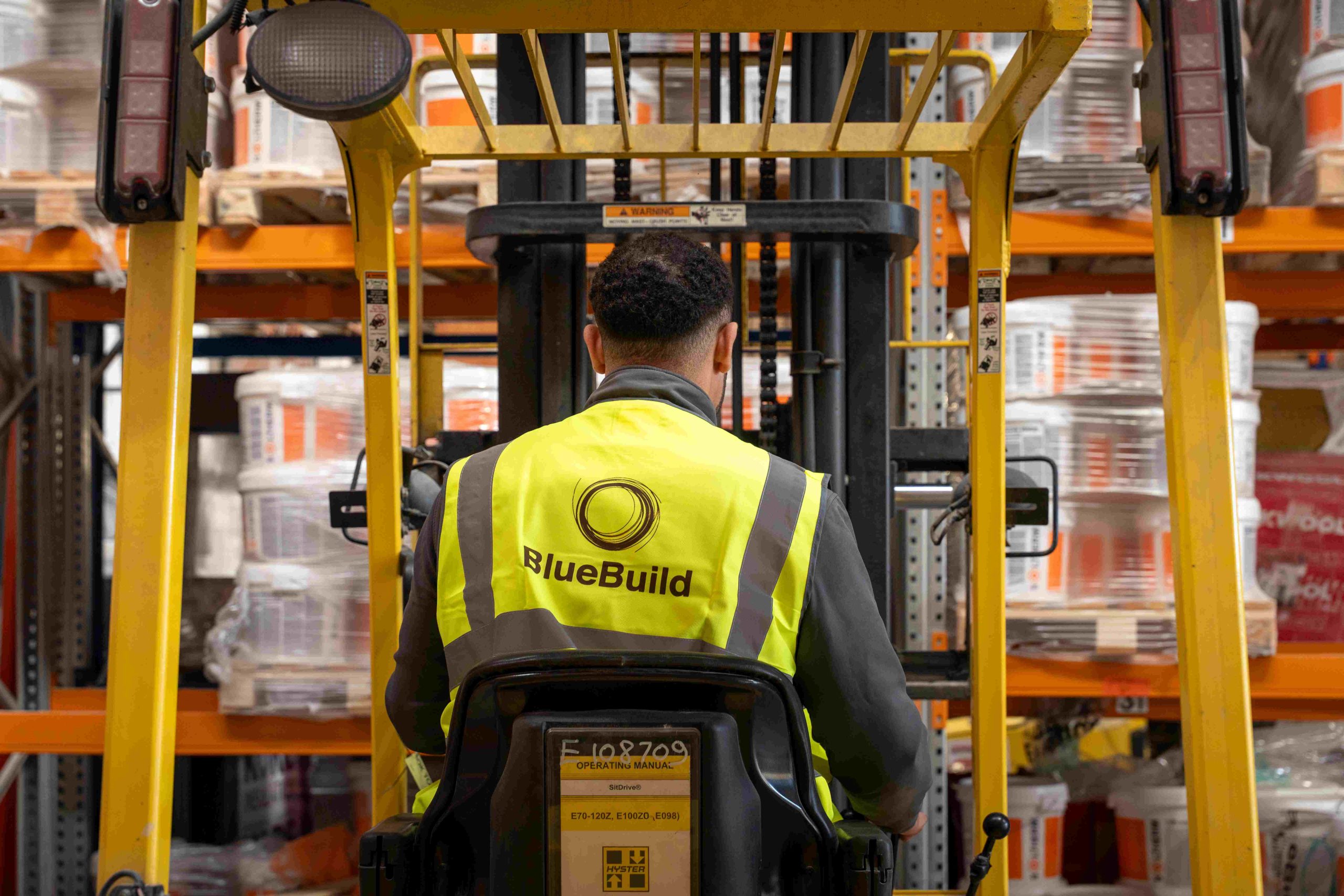 A rear view of a warehouse worker wearing a high-visibility BlueBuild vest, operating a yellow forklift machine inside a large storage facility with tall inventory shelving loaded with building materials.