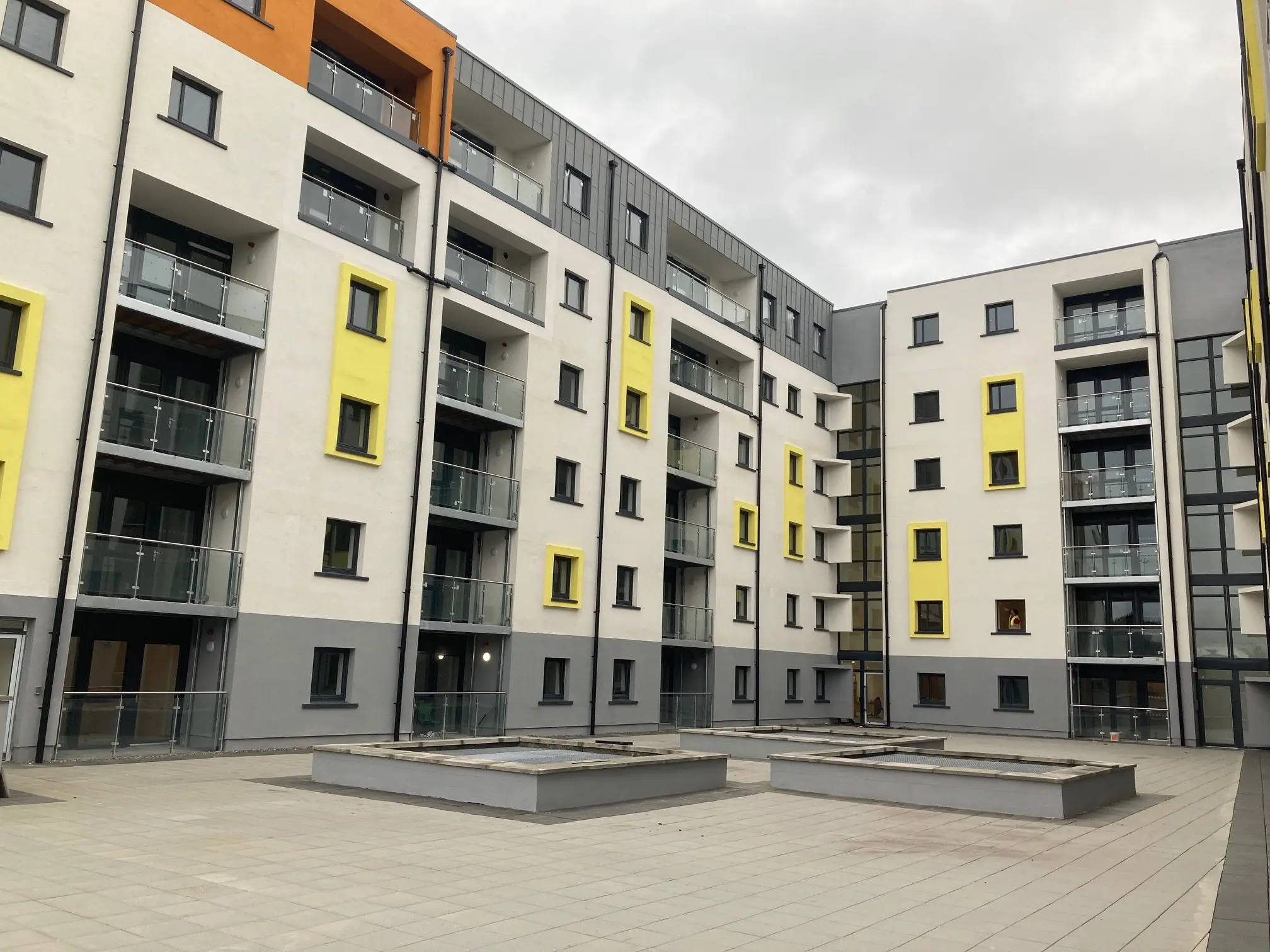 Exterior view of a modern multi-story apartment block complex with white, grey, yellow, and orange architectural accents, surrounding a paved central courtyard.