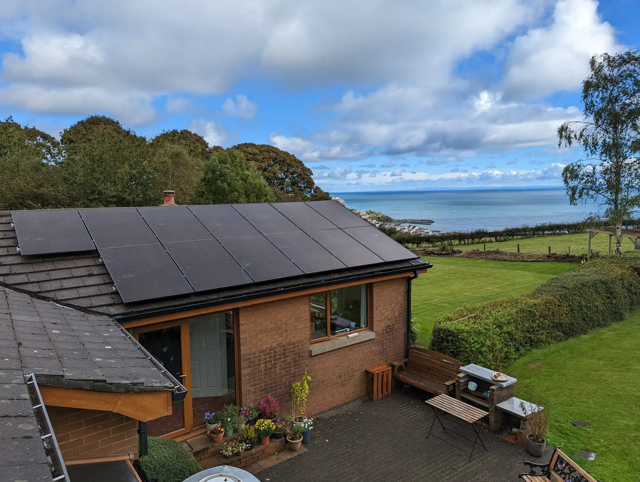 Black solar panels installed across a tiled roof of a brick house, overlooking a paved patio, a large green lawn, and a scenic coastal ocean view.