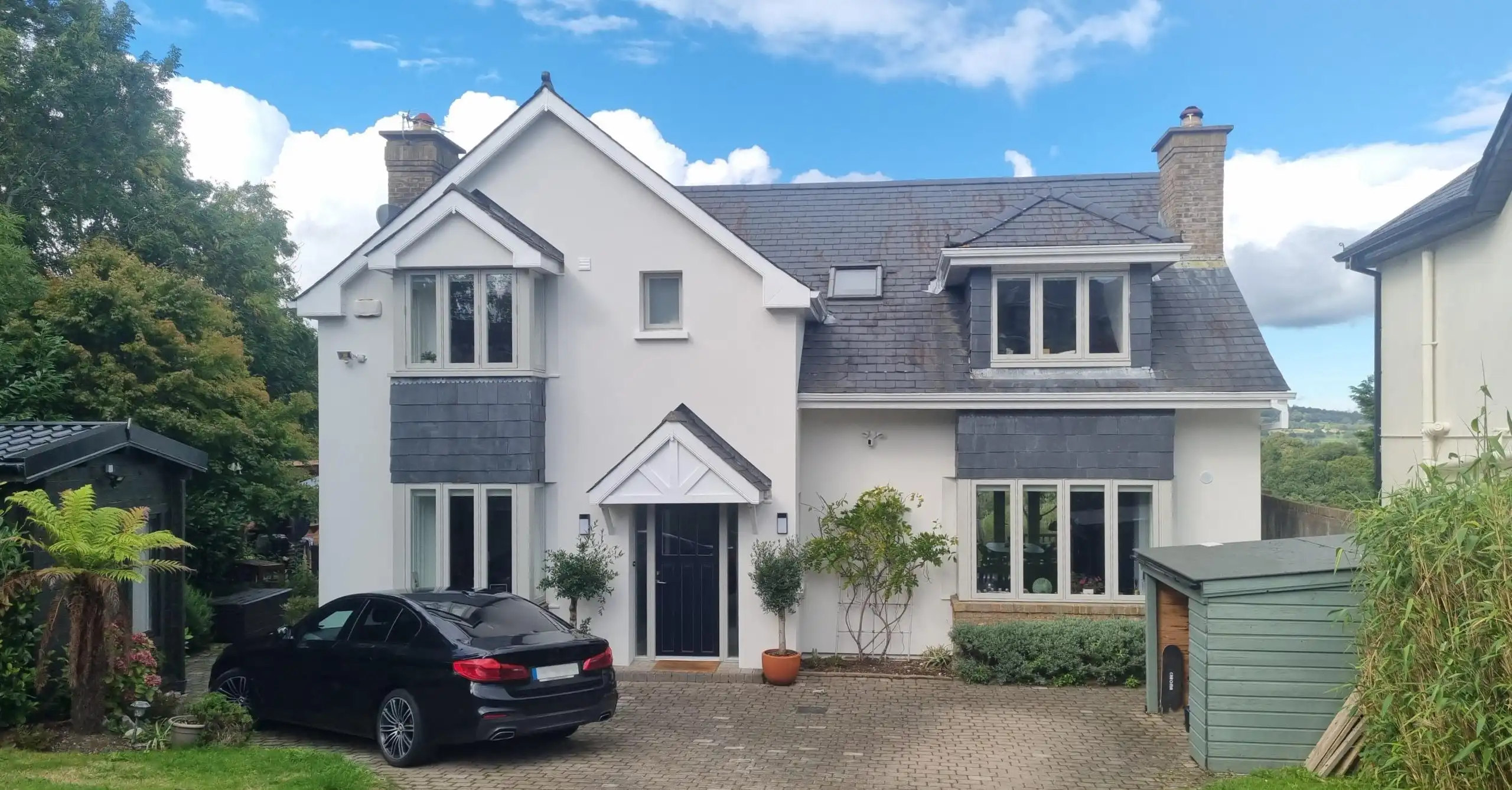 A two-story detached house with a white rendered facade, dark grey slate detailing, and a black front door, featuring a block-paved driveway with a parked black car and a small garden shed.